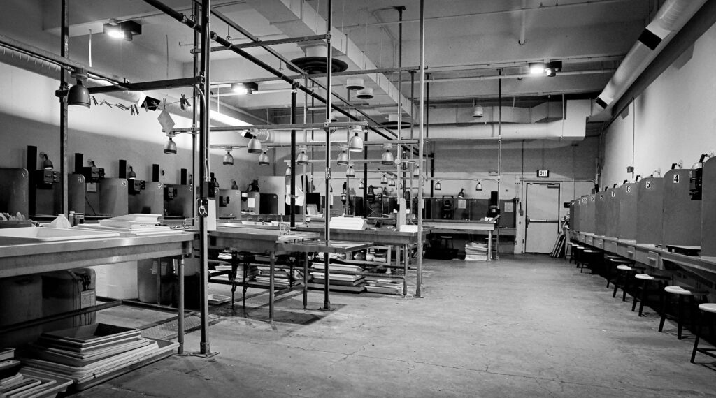 The black and white darkroom with the lights on: enlarging stations along the walls and a long processing sink